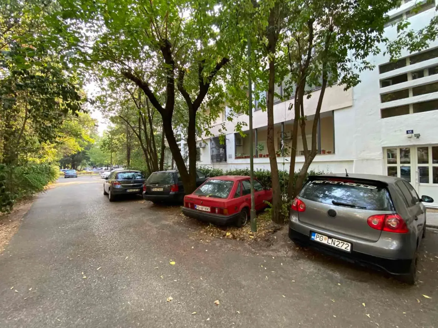 Eksterijer stambene zgrade sa parkingom i drvećem.