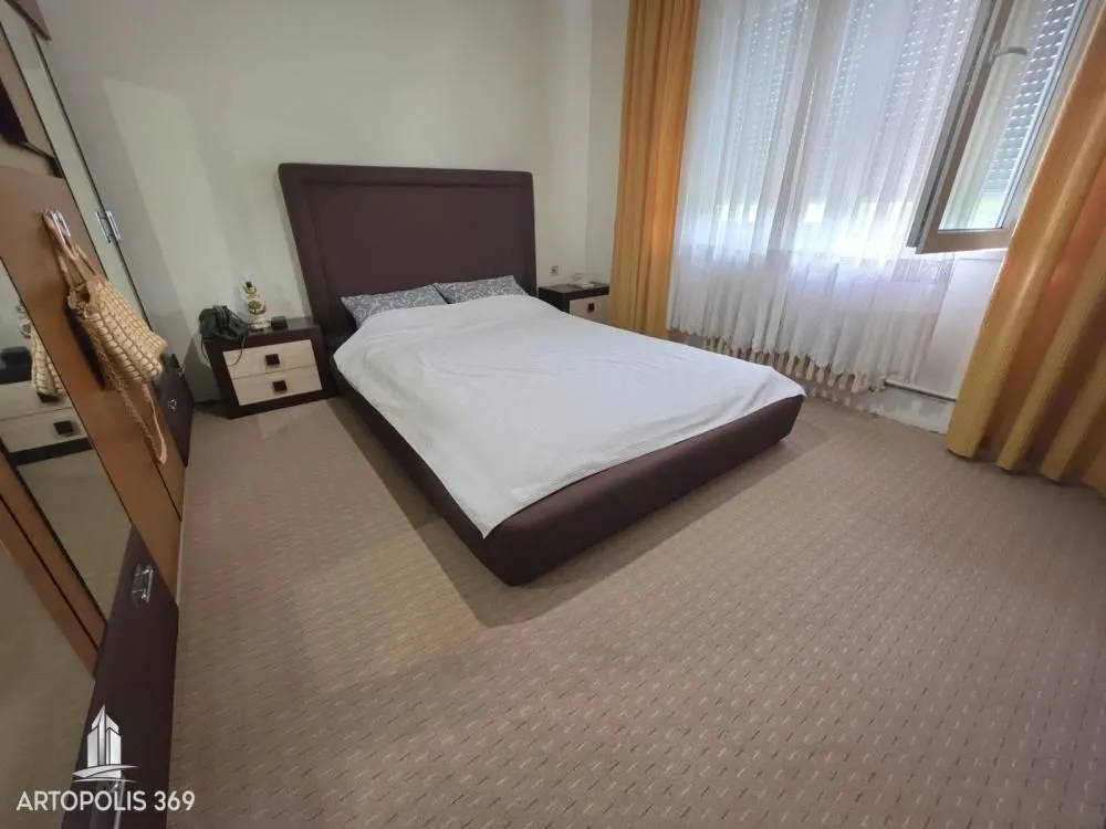 Nameštena spavaća soba sa bračnim krevetom, noćnim stočićima i prozorom sa zavesama.