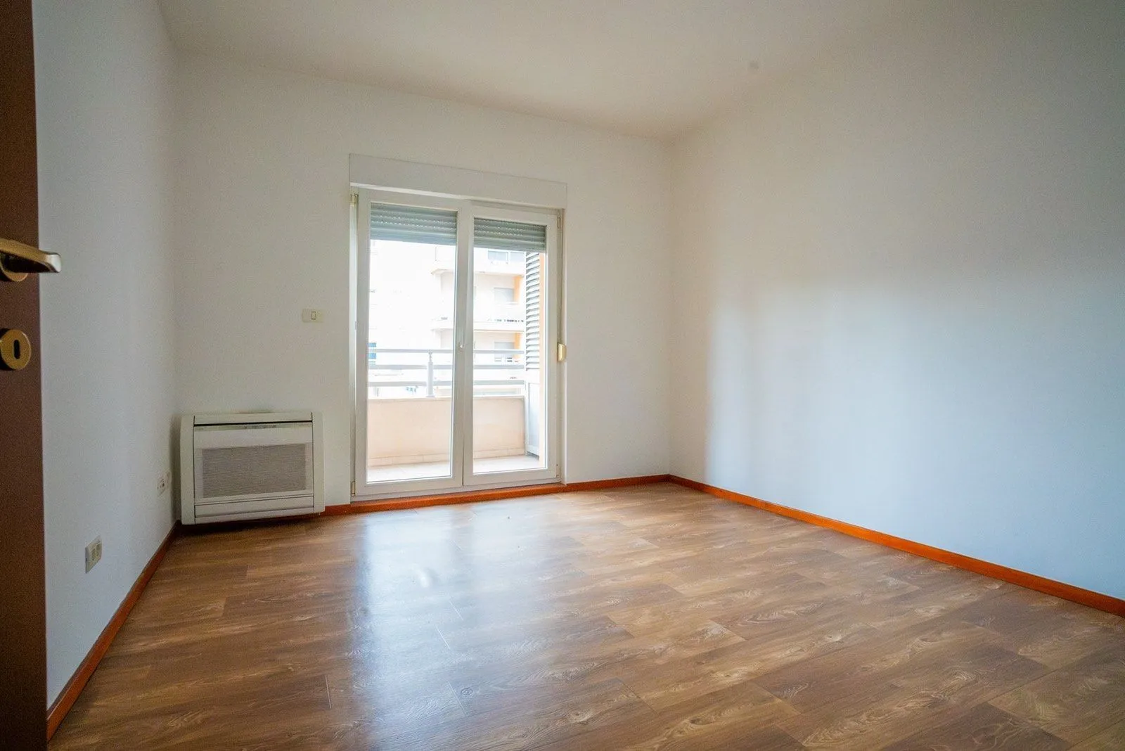 Prazna soba sa svijetlim zidovima, parketom i izlazom na balkon.