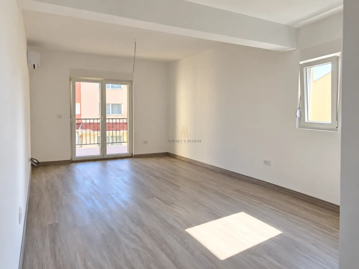 Prazna soba sa laminatom, bijelim zidovima i izlazom na balkon.