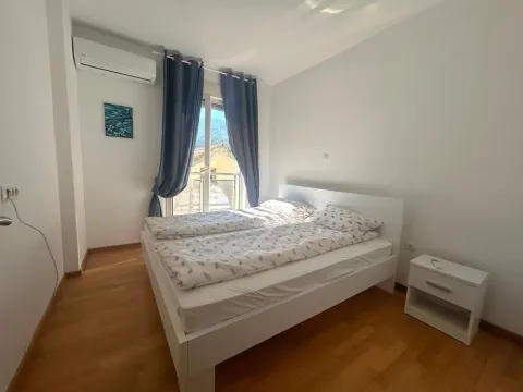 Svjetla spavaća soba sa bračnim krevetom, bijelim noćnim stočićem i prozorom sa plavim zavjesama. - image 7