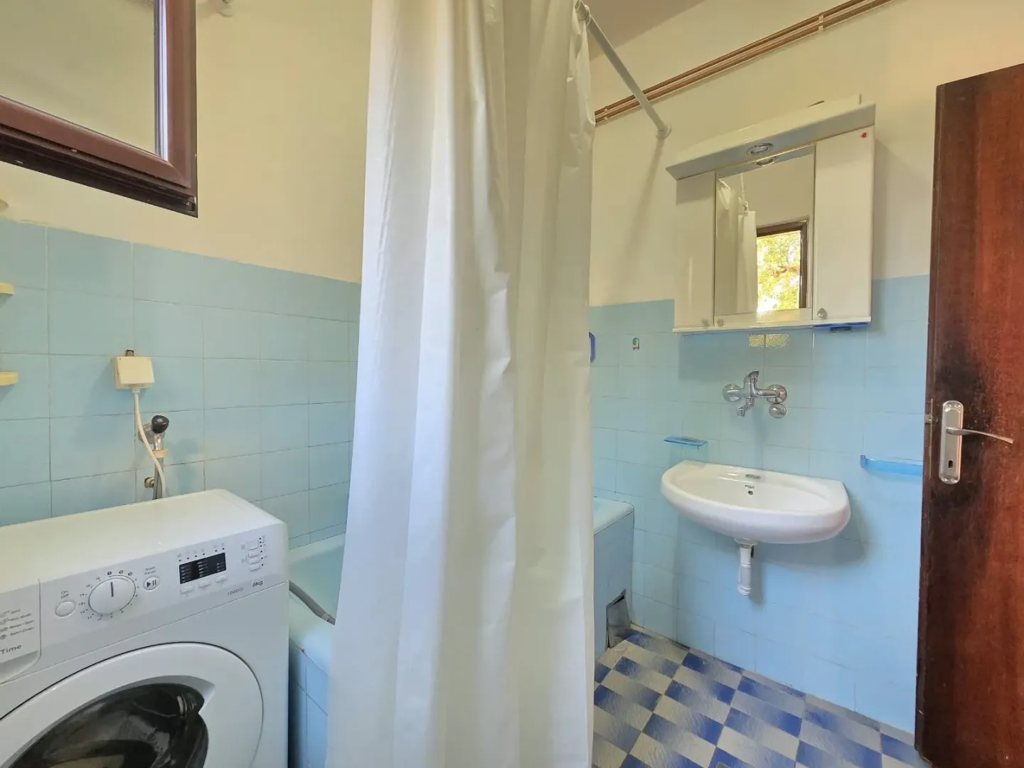 Kupatilo sa plavim pločicama, umivaonikom, kadom i veš mašinom.