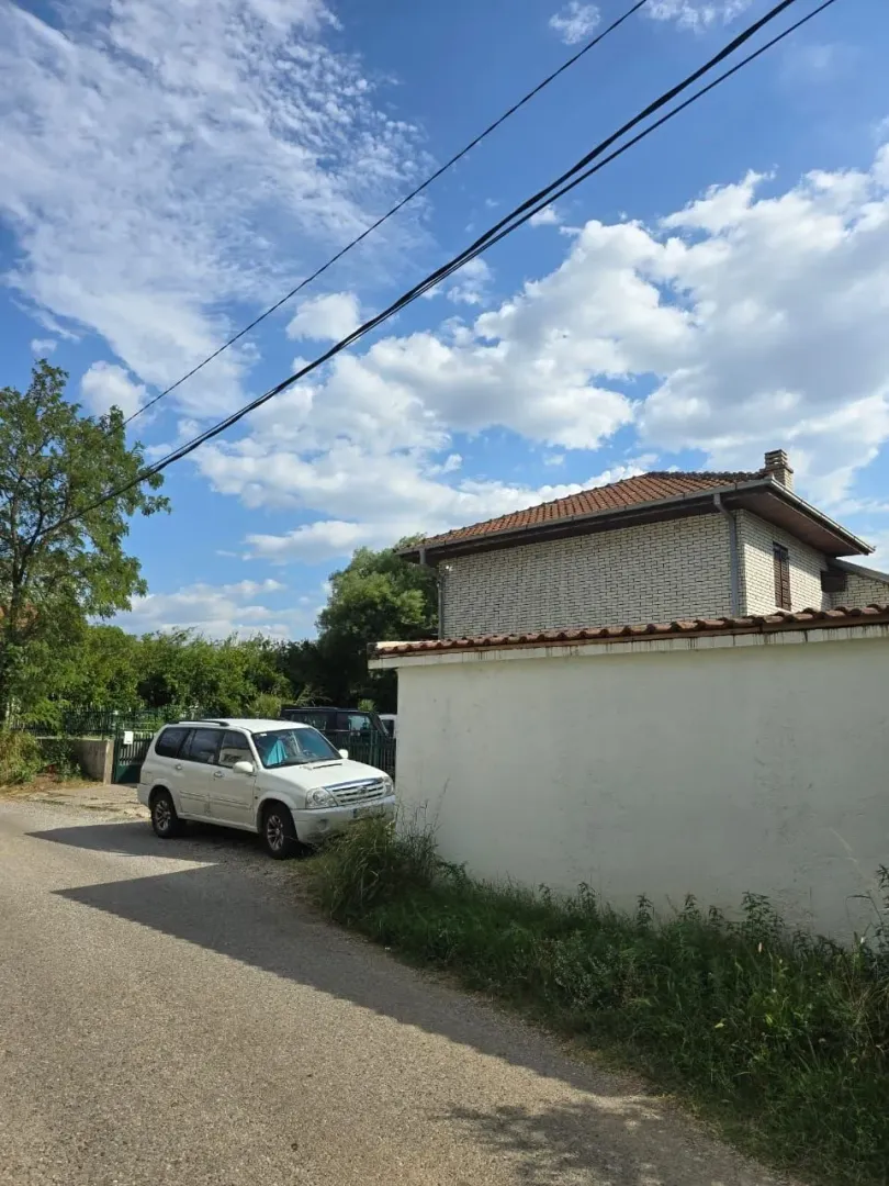 Spoljašnji izgled dvospratne kuće sa dvorišnim zidom i automobilom parkiranim pored puta.