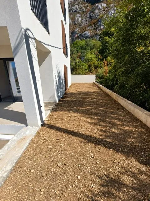 Dvorište sa šljunkom pored moderne zgrade i zelenog brda u pozadini.