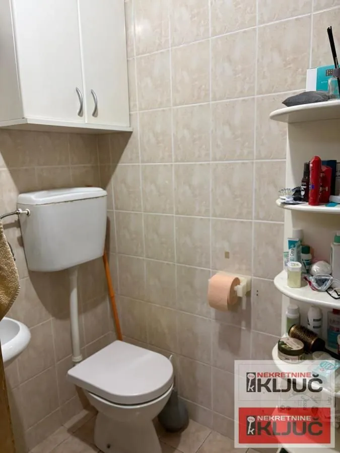 Manje kupatilo sa wc šoljom, belim ormarićem i zidnim pločicama.