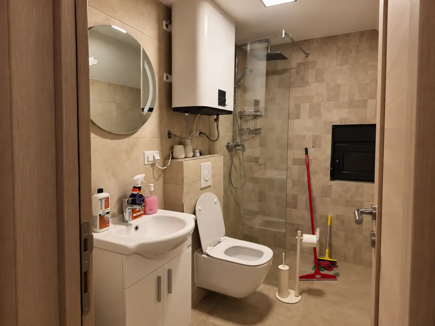 Manje kupatilo sa tuš kabinom, umivaonikom i wc šoljom obloženo bež pločicama.