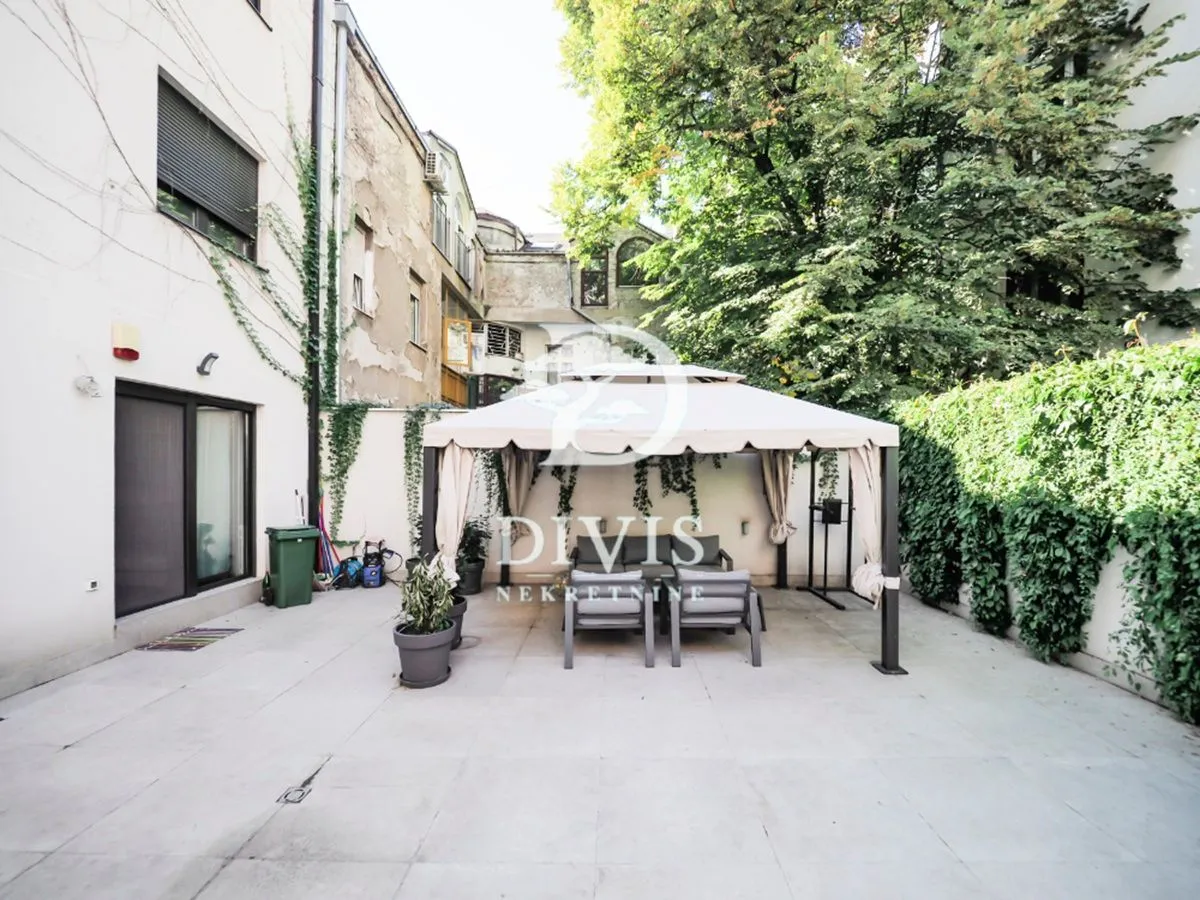 Paved courtyard with seating furniture under a canopy and greenery.