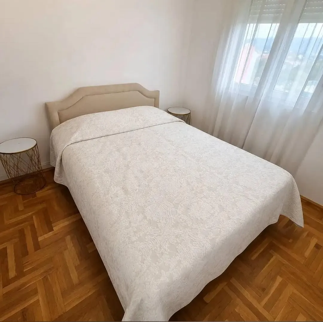 Svijetla spavaća soba sa bračnim krevetom, parketom i prozorom sa zavjesama.
