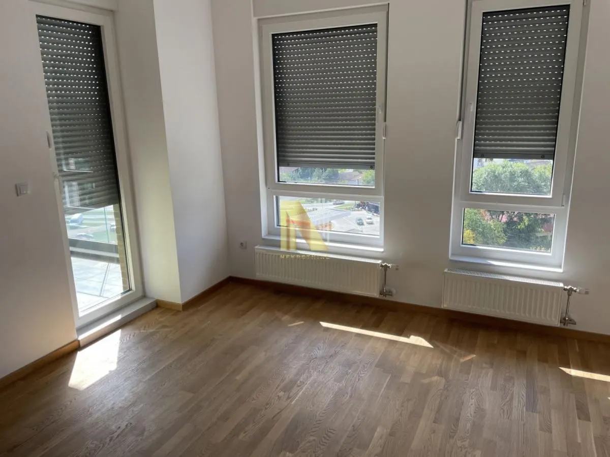 Empty room with wooden flooring, white walls, and large windows with roller shutters.