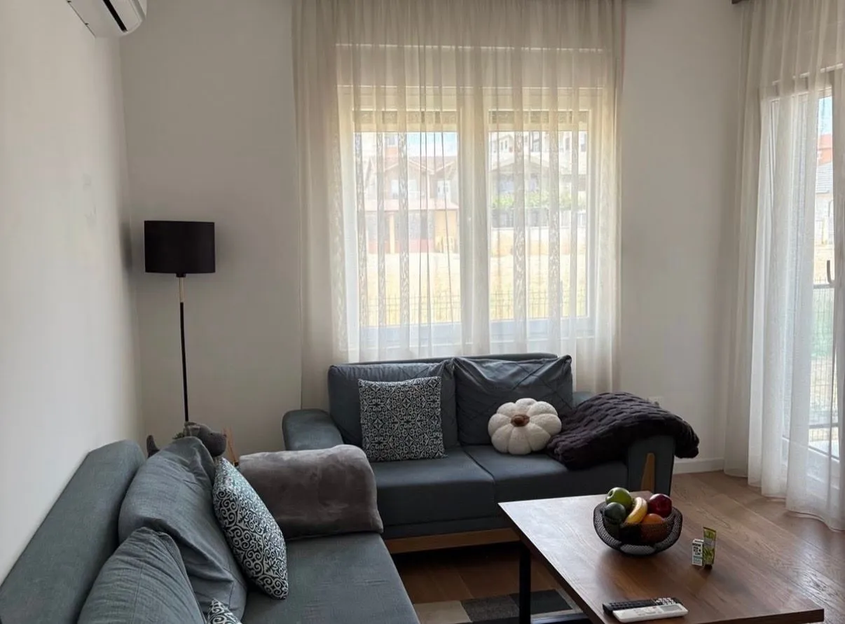 Dnevna soba sa garniturom, stočićem i prozorima sa svijetlim zavjesama.