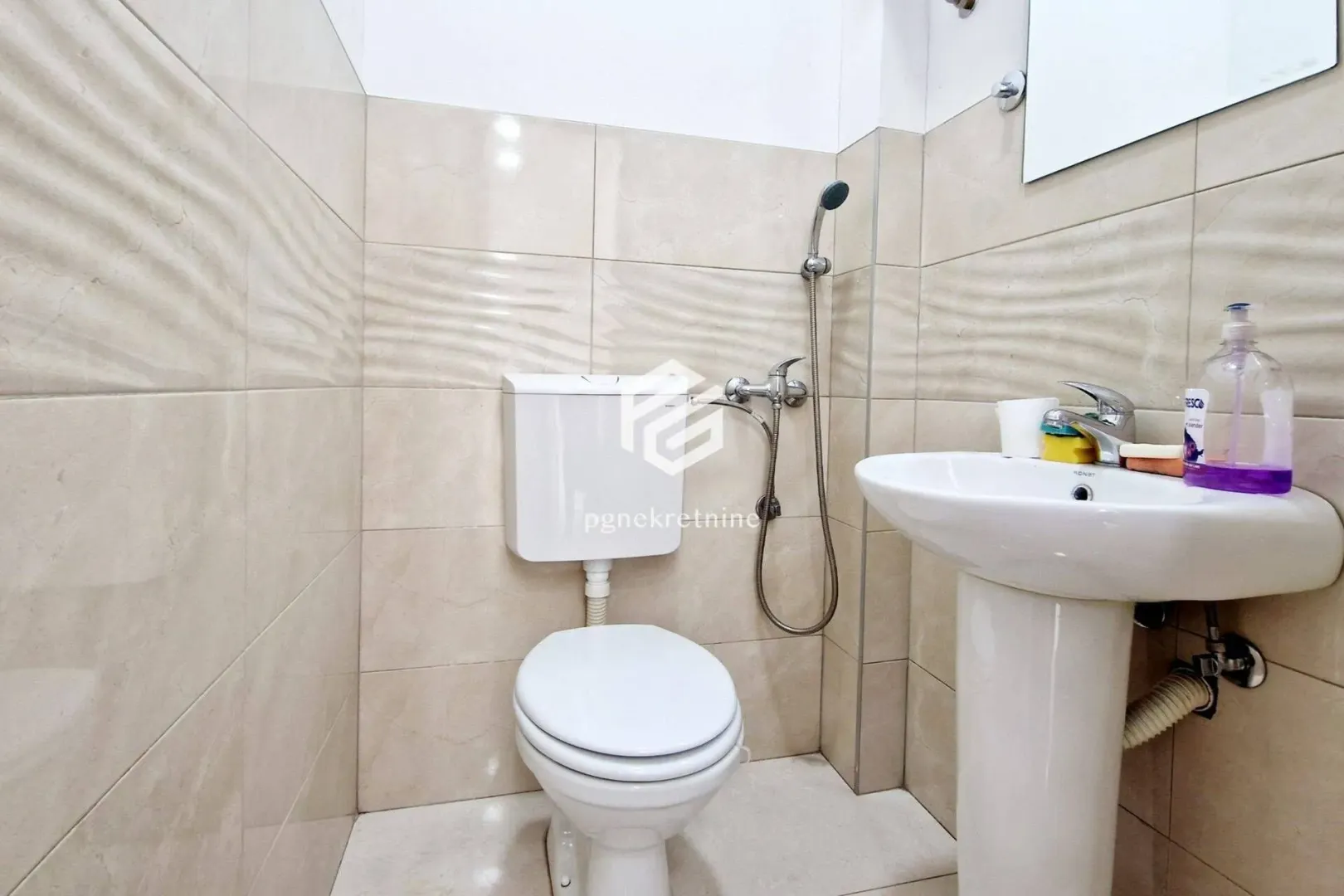 Smaller bathroom lined with beige tiles featuring a toilet and washbasin.