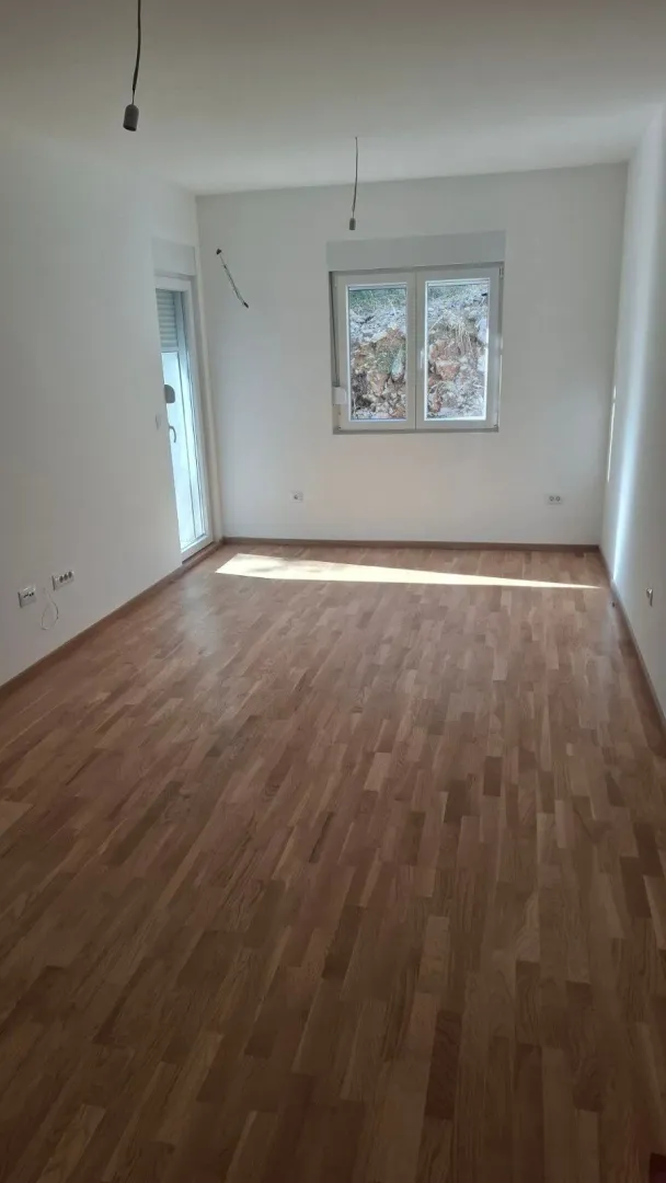 Prazna neopremljena prostorija sa drvenim podom, bijelim zidovima i prozorom.