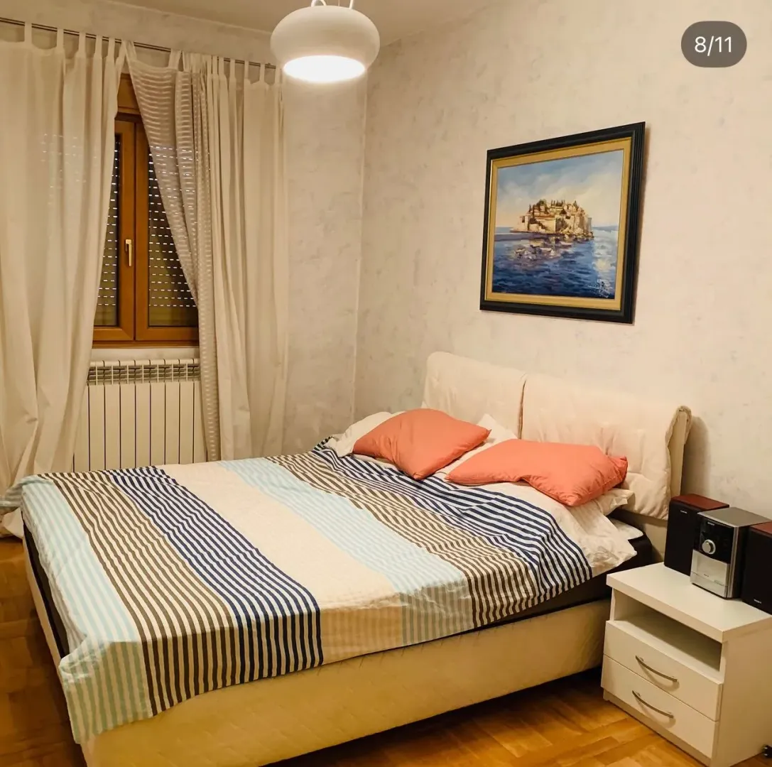 Uređena spavaća soba sa bračnim krevetom, bijelim zavjesama i umjetničkom slikom na zidu.