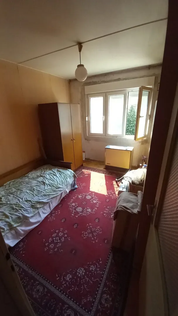 Skromno opremljena spavaća soba sa krevetom, ormarom i crvenim tepihom.