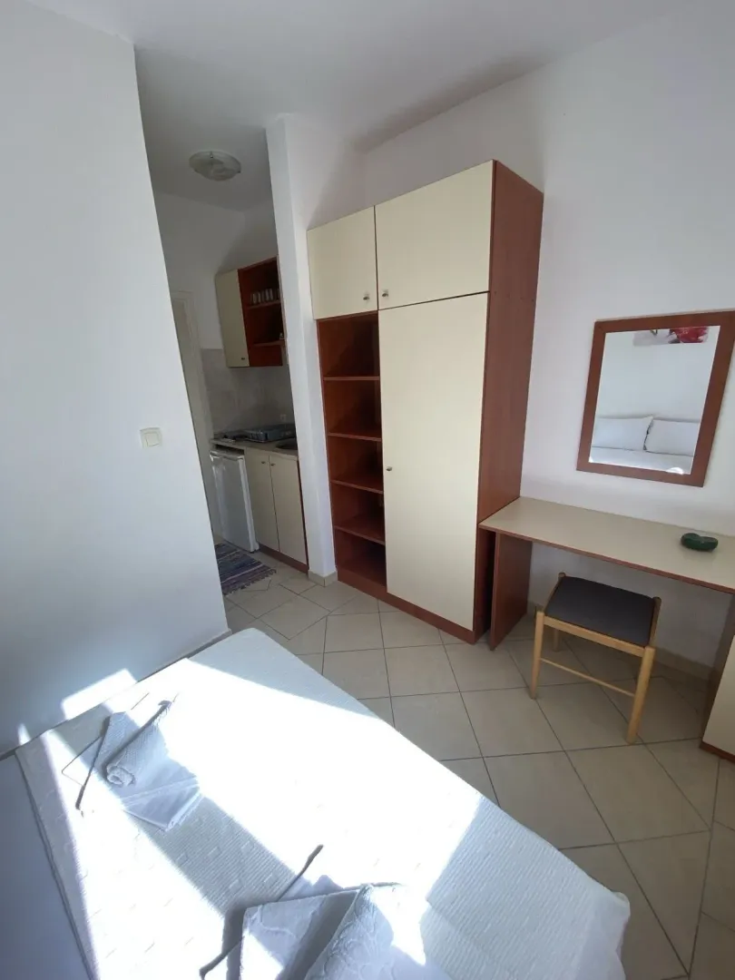 Svijetla spavaća soba sa ormarom, radnim stolom i kuhinjom u pozadini.