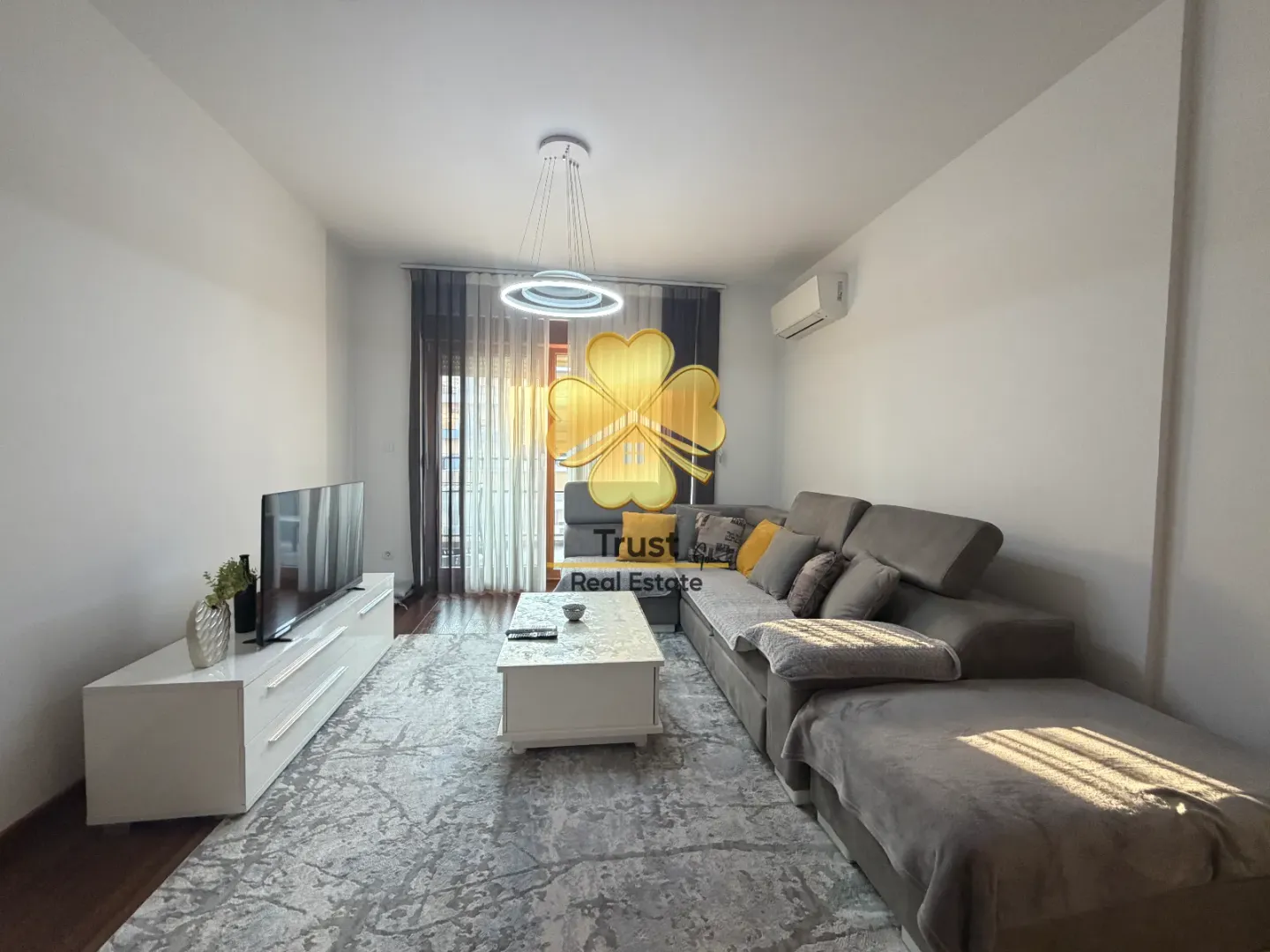 Prostrana dnevna soba sa ugaonom garniturom, TV komodom i modernim lusterom.