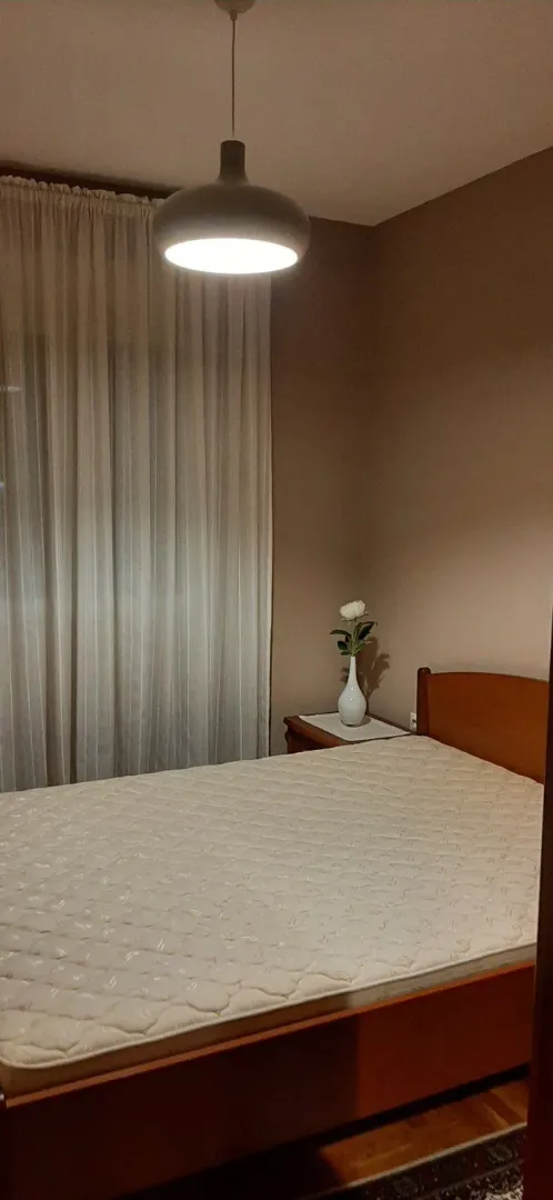 Spavaća soba sa bračnim krevetom, noćnim stočićem i zavjesama na prozoru.