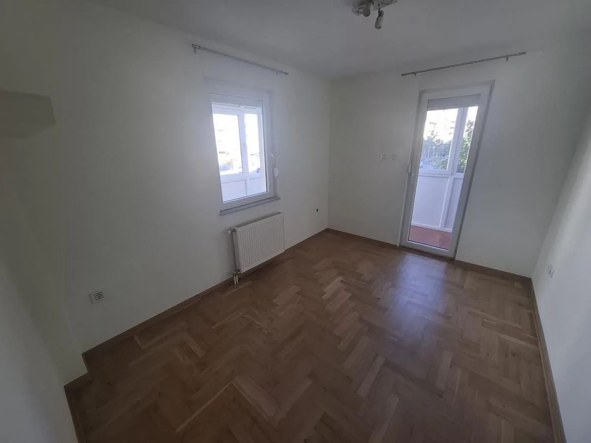 Prazna soba sa parketom, prozorom i balkonskim vratima.