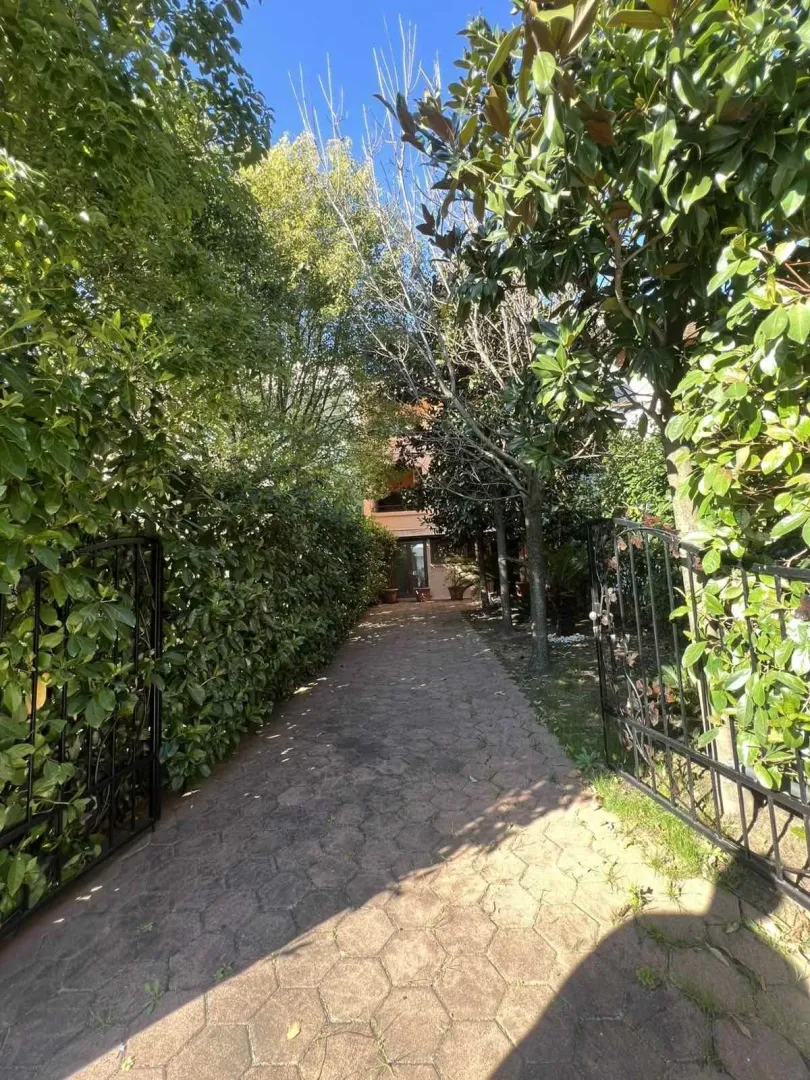 Paved driveway leading to the house entrance framed by an open iron gate and lush greenery.