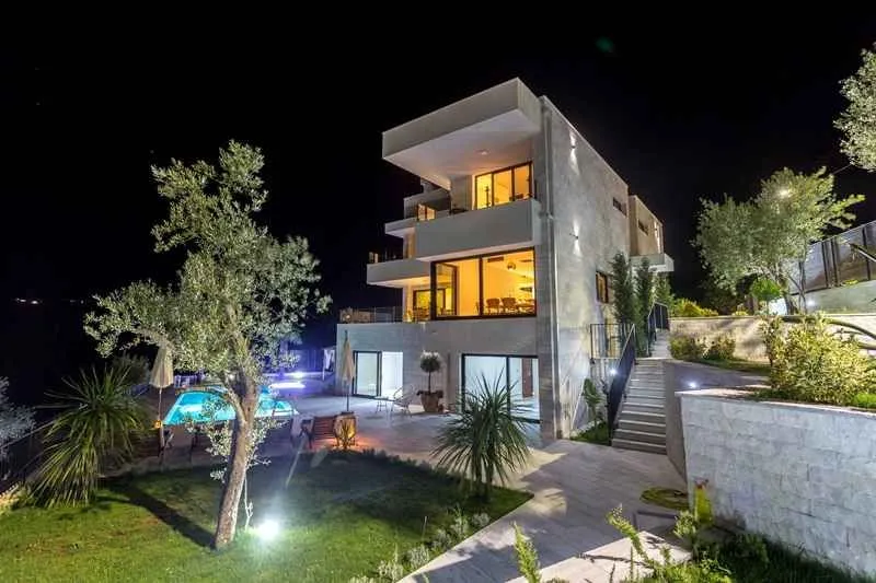 Noćni prikaz moderne višespratne kuće sa bazenom i uređenim dvorištem.