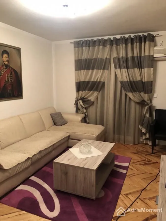 Dnevna soba sa ugaonom garniturom, stočićem i prozorom sa zavesama.