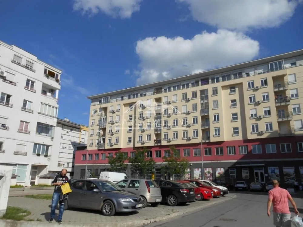 Pogled na spoljašnjost stambene zgrade sa parkingom ispred.