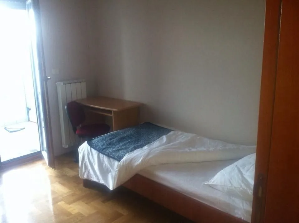 Jednostavna spavaća soba sa jednokrevetnim ležajem, radnim stolom i izlazom na balkon.