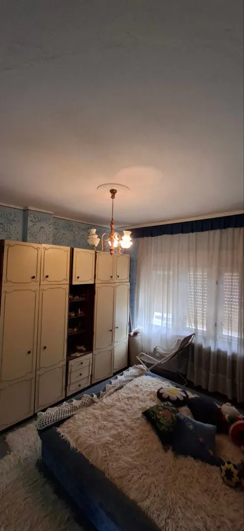 Spavaća soba sa velikim ormarom, krevetom i prozorom sa belim zavesama.