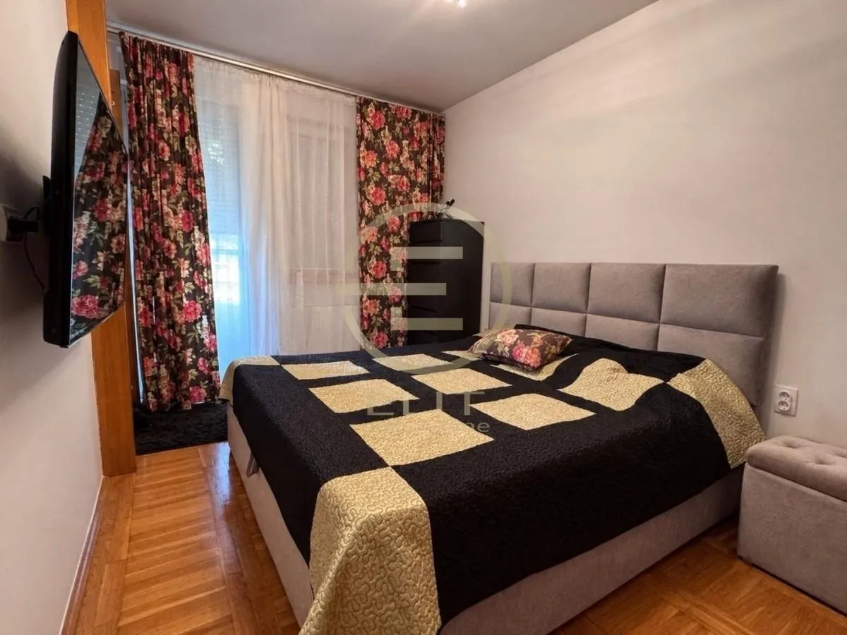 Nameštena spavaća soba sa bračnim krevetom, TV-om i prozorom sa zavesama.