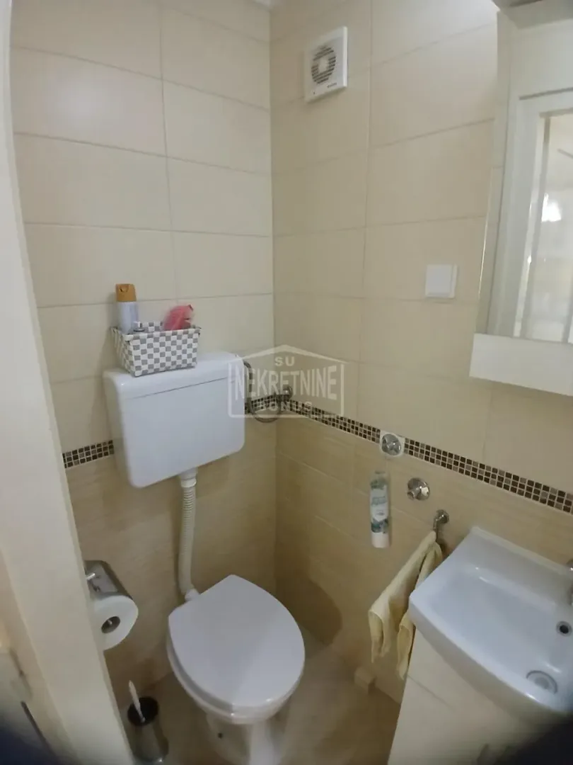 Manje kupatilo sa WC šoljom, umivaonikom i bež pločicama.