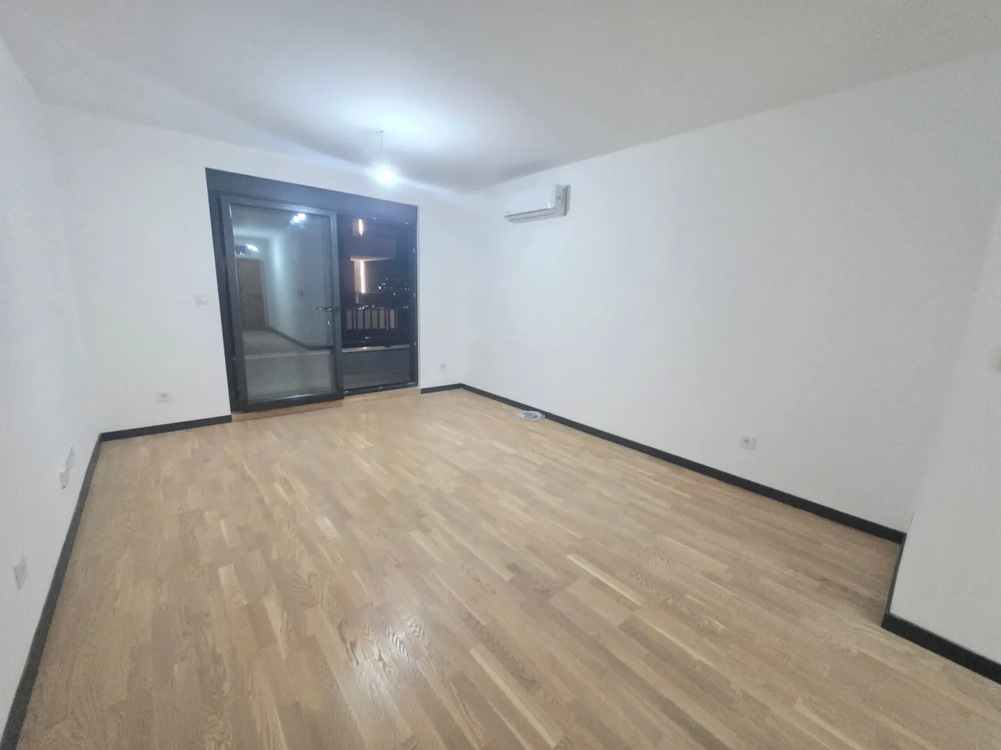 Prazna soba sa drvenim parketom, bijelim zidovima i klima uređajem.
