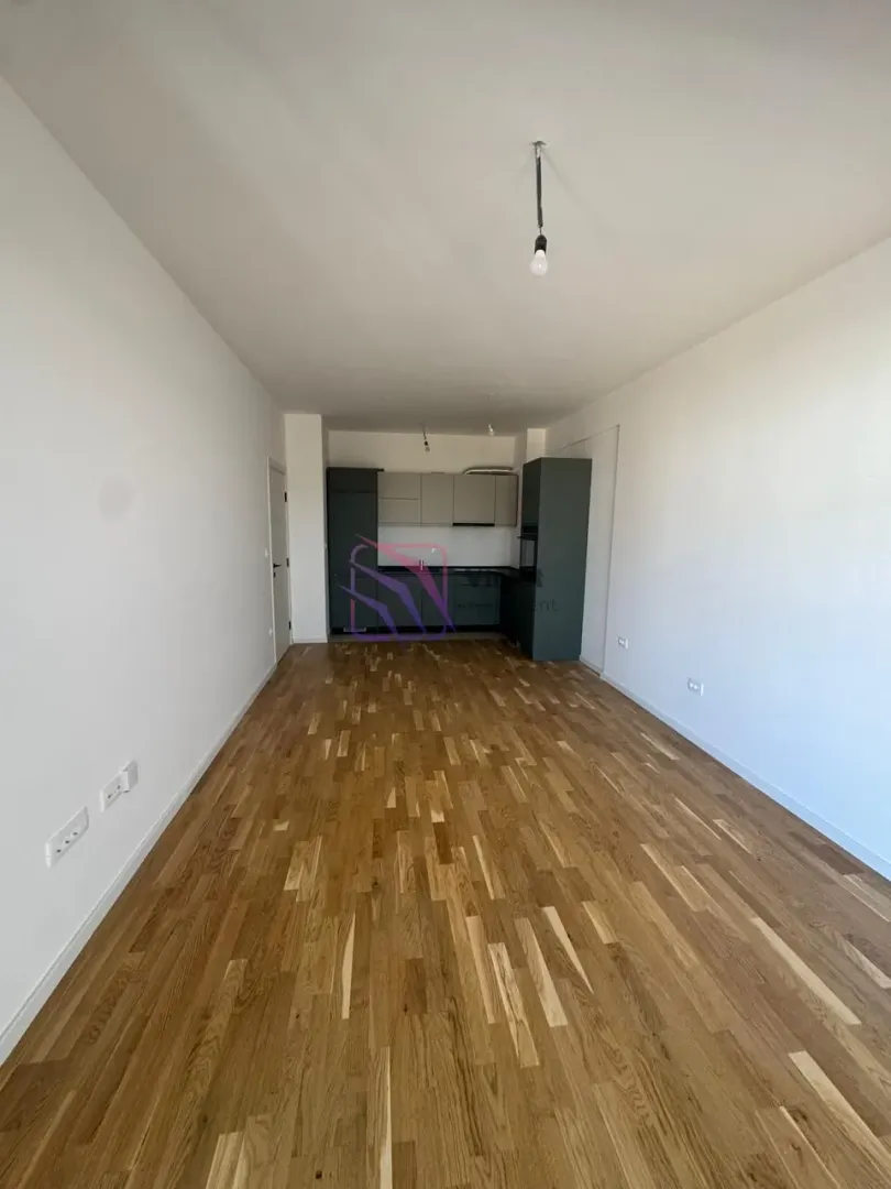 Prazna dnevna soba sa drvenim podom i pogledom na kuhinju u pozadini.