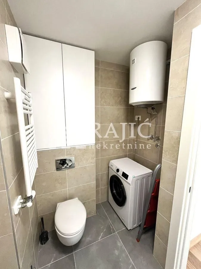 Manje kupatilo sa veš mašinom, wc šoljom i ugradnim ormarićem.