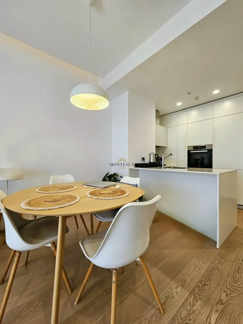 Dining area with a wooden table and chairs in a modern apartment with the kitchen in the background.