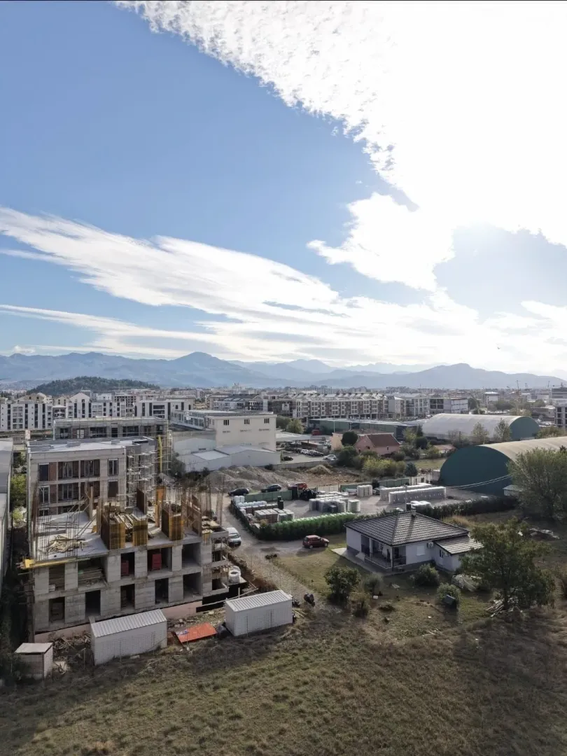 Pogled iz vazduha na gradilište i okolne objekte sa planinama u pozadini.