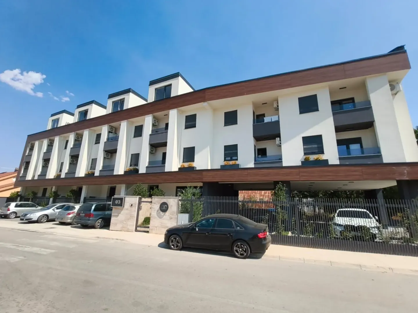 Pogled na spoljašnjost moderne stambene zgrade sa parkingom i ograđenim dvorištem.