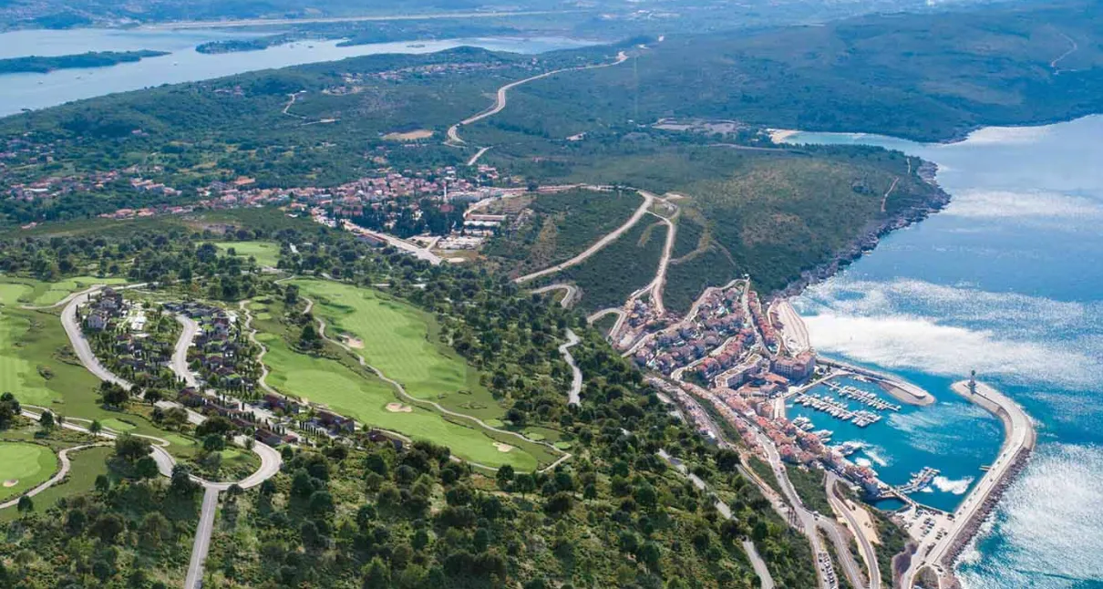 Pogled iz vazduha na obalu, naselje i marinu sa zelenim površinama.