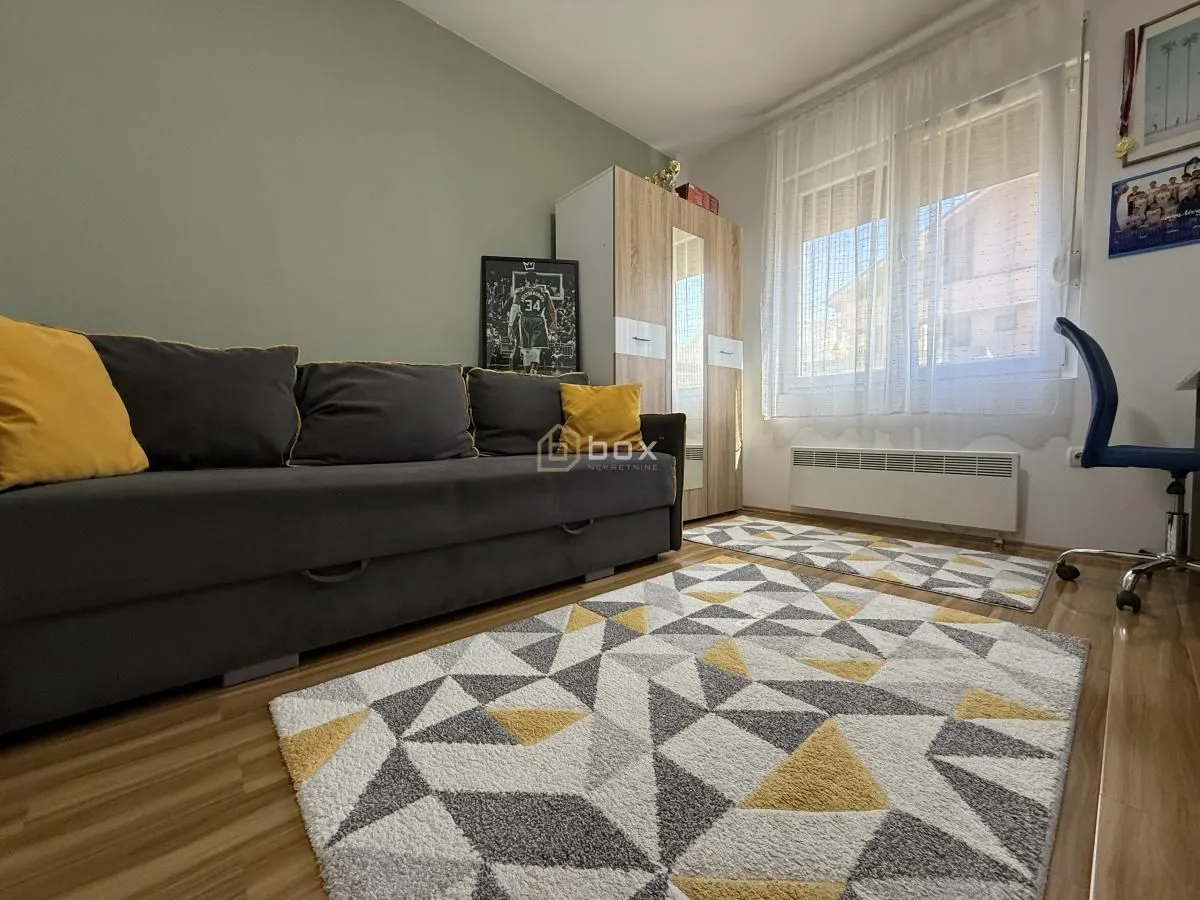 Dnevna soba sa garniturom za sedenje, ormarom i prozorom sa belim zavesama.