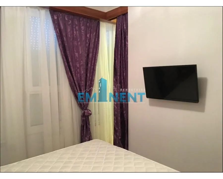 Bedroom with a bed, a window with purple curtains, and a wall-mounted TV.