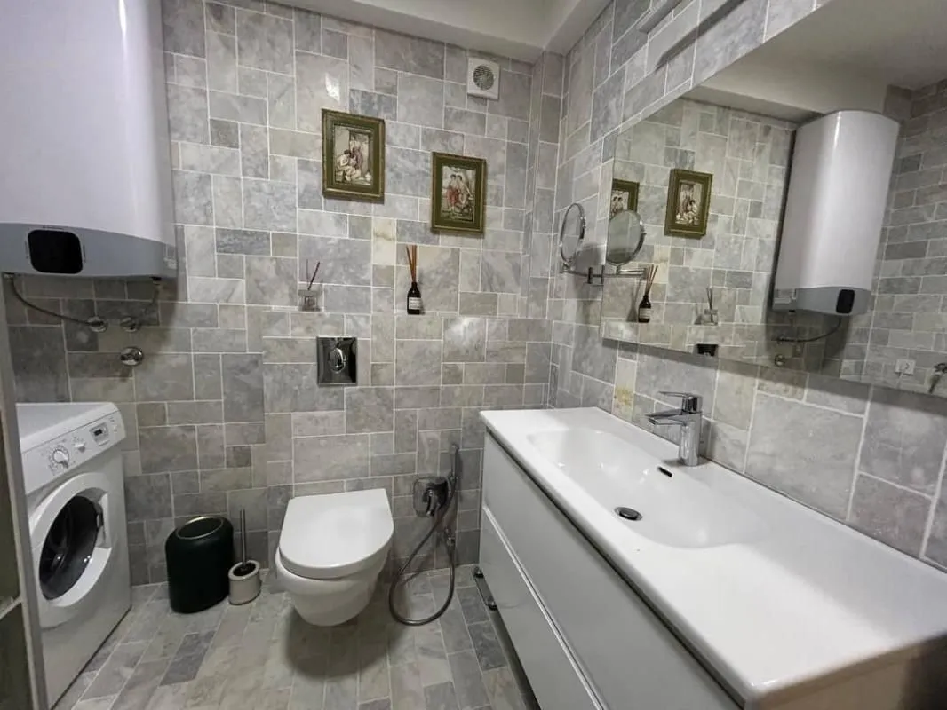 Kupatilo obloženo sivim pločicama sa umivaonikom, WC šoljom, veš mašinom i bojlerom.