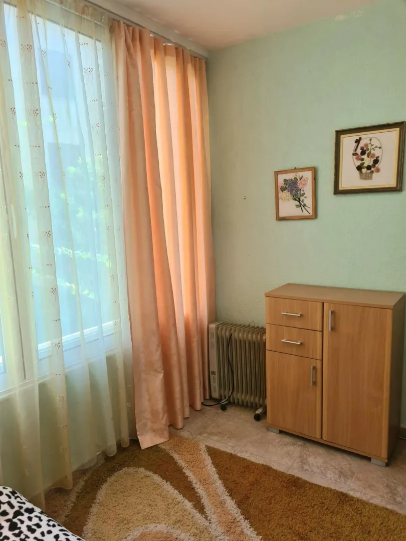 Manja soba sa drvenom komodom, radijatorom i prozorom sa zavjesama.