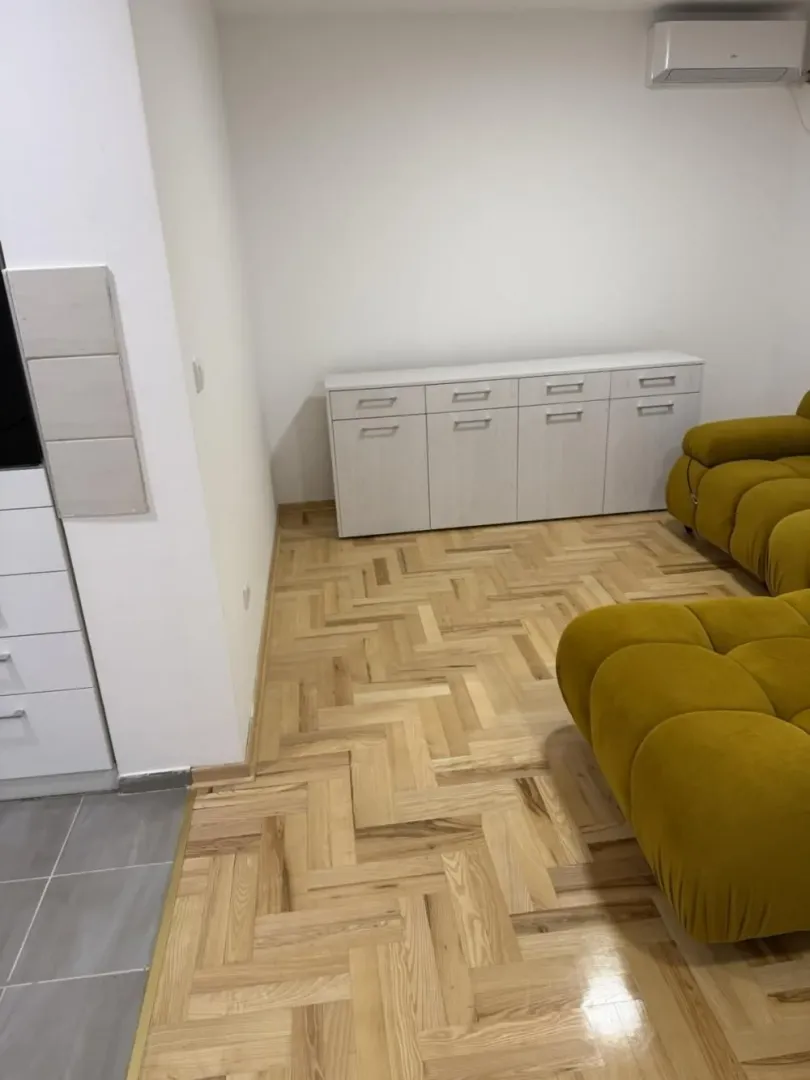 Deo dnevne sobe sa parketom, belom komodom i delom žute garniture.
