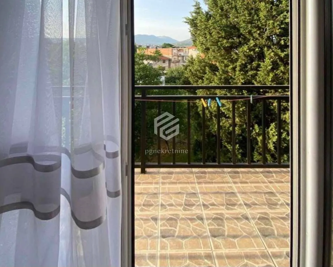 Pogled kroz balkonska vrata na popločanu terasu i zelenilo.