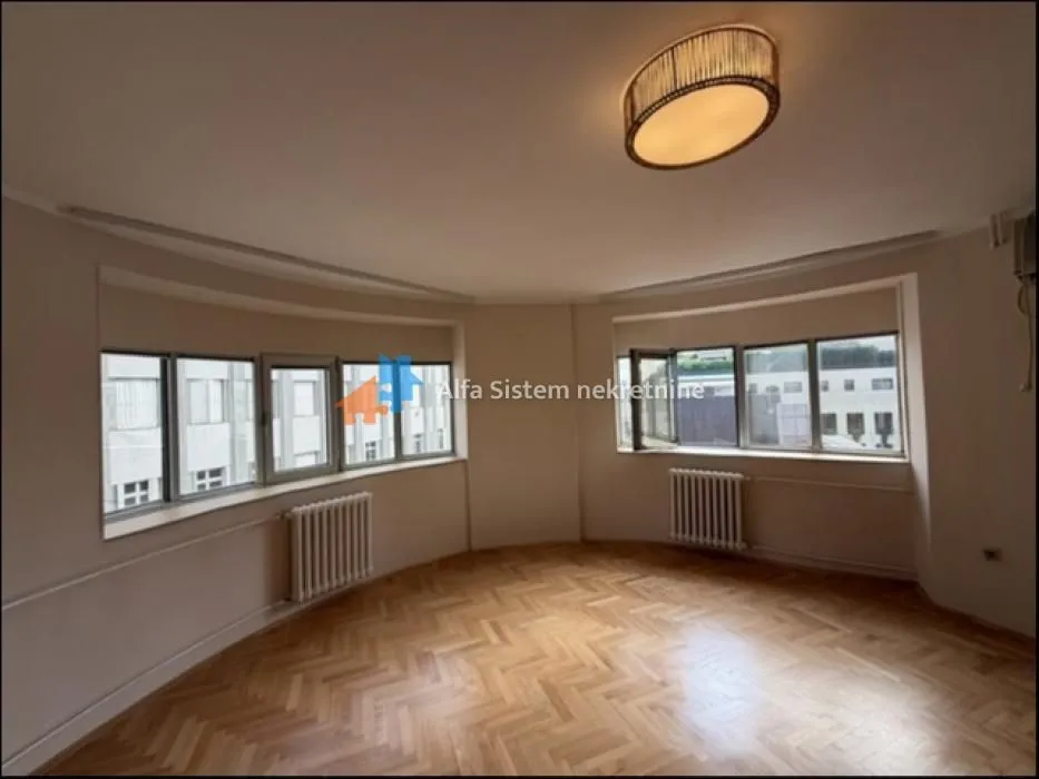 Empty living room with parquet flooring and large windows.