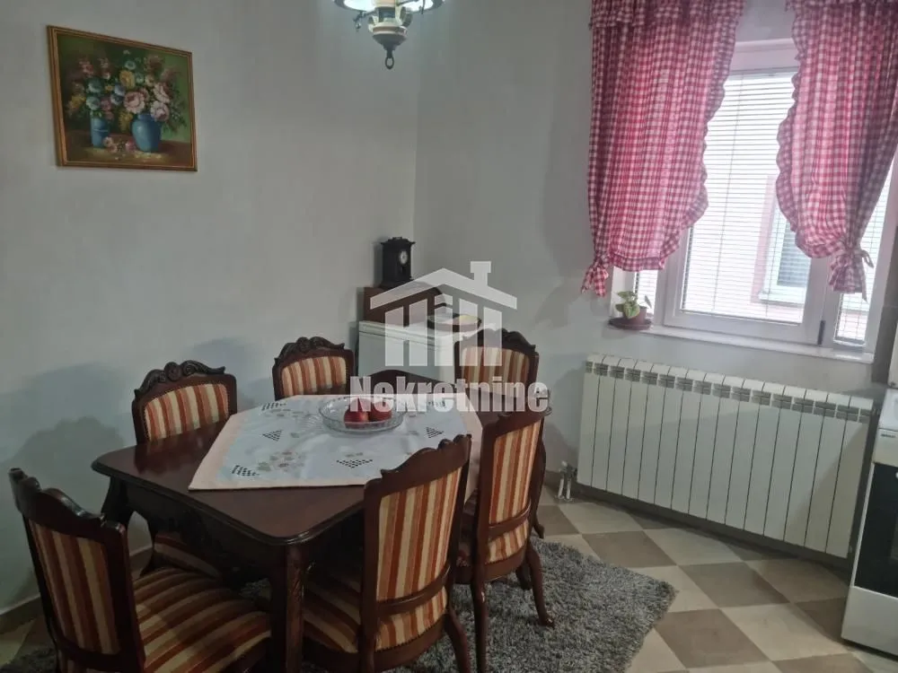 Trpezarija sa drvenim stolom i stolicama, smeštena pored prozora.