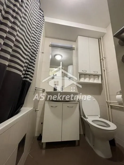 Manje kupatilo sa umivaonikom, WC šoljom, ormarićem i kadom sa zavesom.