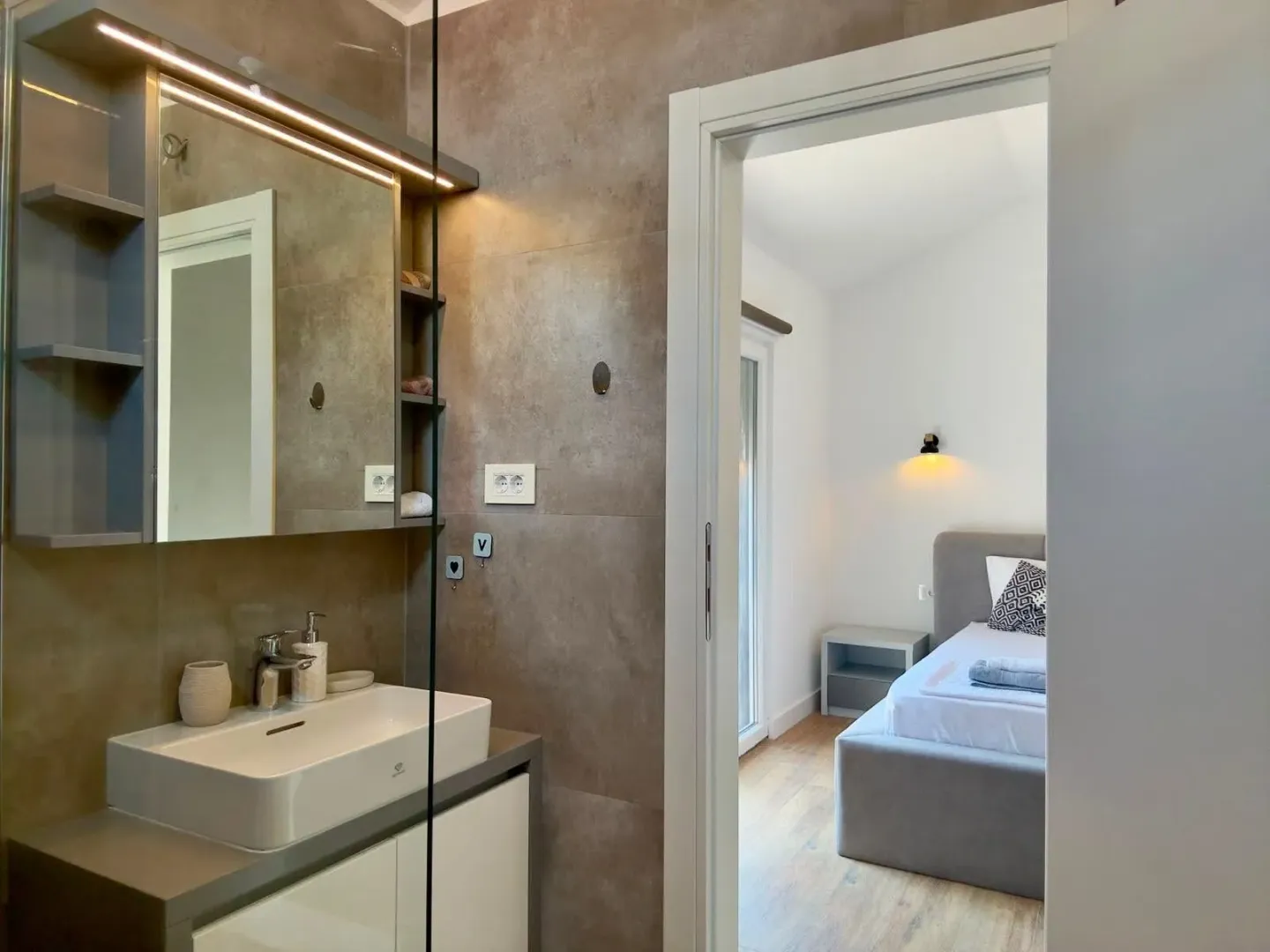 Modern bathroom with a sink and mirror, overlooking the bedroom through an open door.