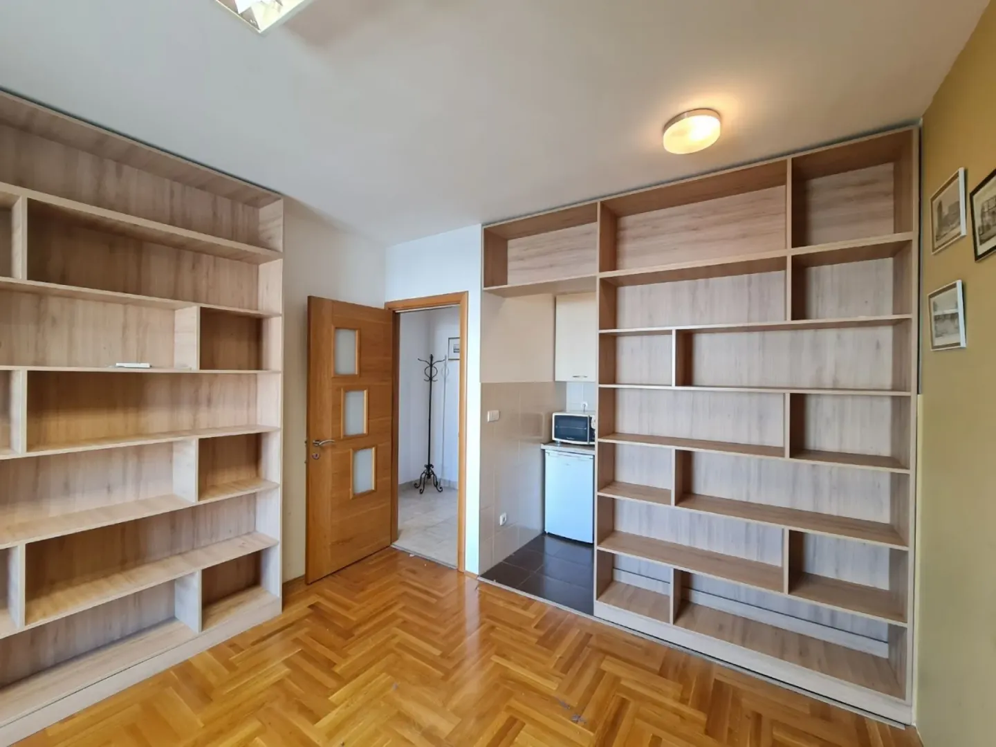 Bright room with parquet flooring, built-in wooden bookshelves, and a view of a kitchenette.