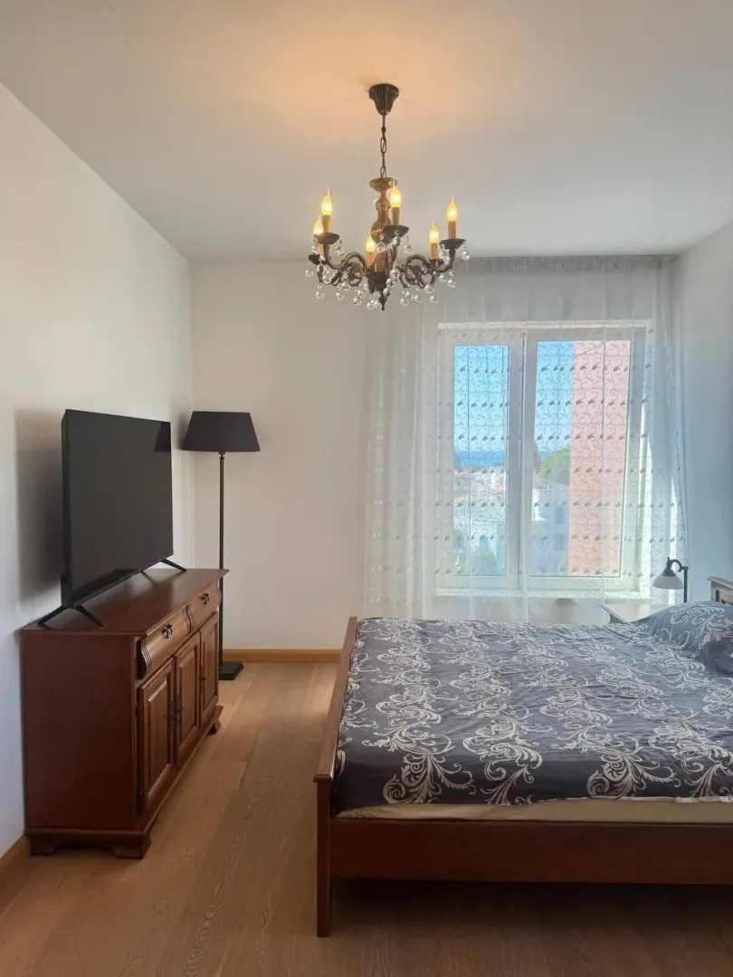 Namještena spavaća soba sa bračnim krevetom, TV komodom i lusterom.