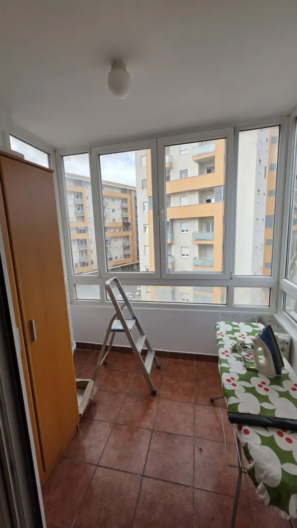 Glazed balcony with floor tiles, an ironing board, and a view of surrounding buildings.